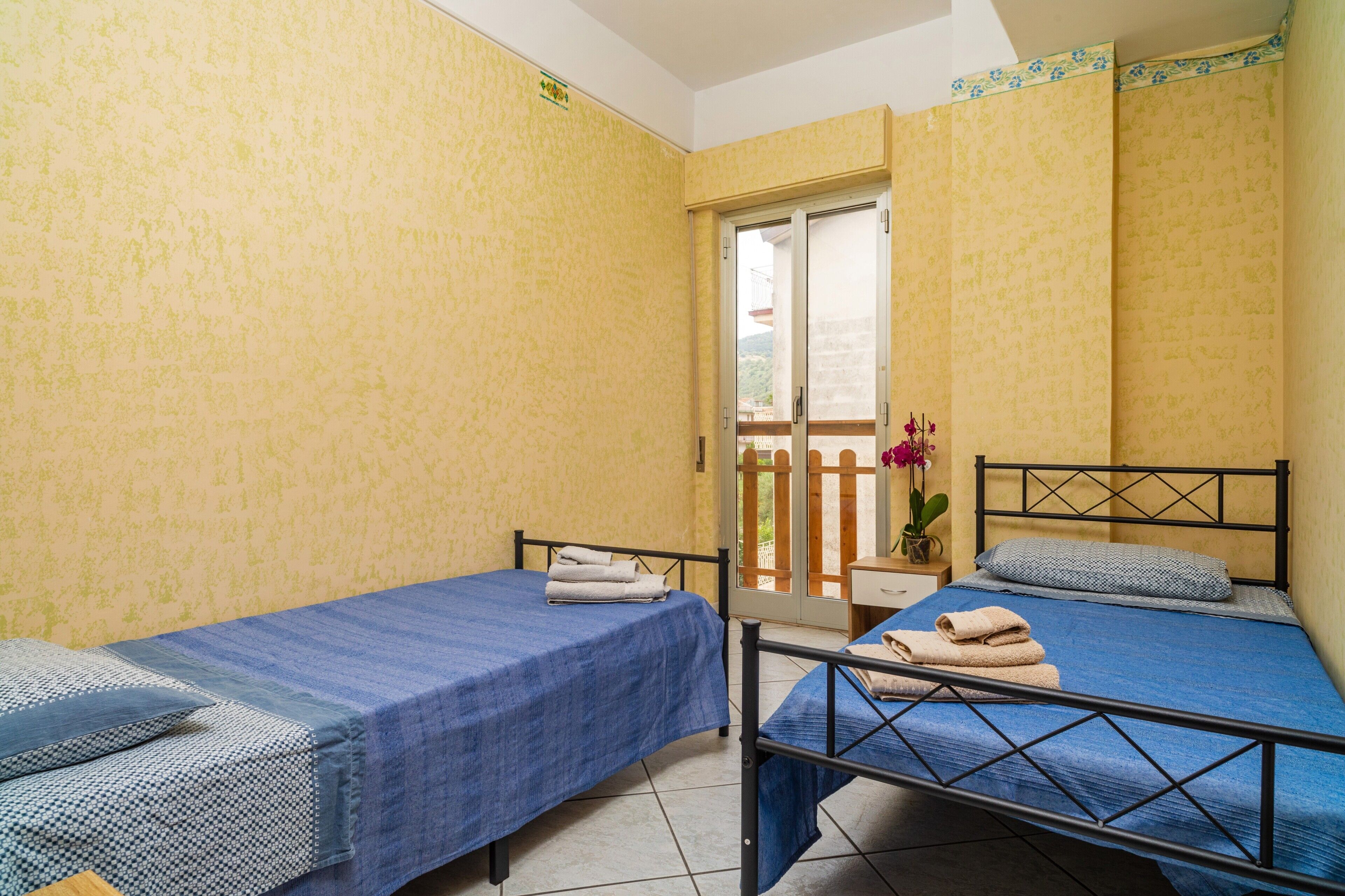 A room with two beds, a small table with a vase of flowers, and a sliding door leading to an outdoor area.