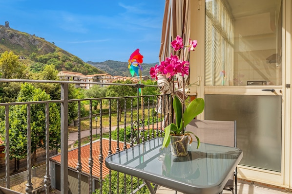 A balcony with a glass table, potted plants, and a view of a hillside with buildings.