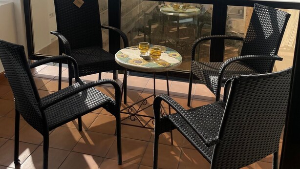 Un balcon avec quatre chaises en osier, une petite table ronde recouverte d’une nappe à motifs floraux et deux verres contenant une boisson jaune.
