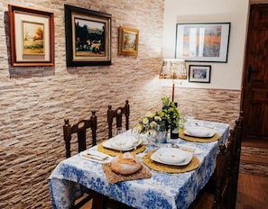 Una mesa de comedor dispuesta con platos, vasos y una hogaza de pan, rodeada de sillas, con cuadros en la pared.