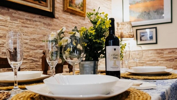 A dining table set with plates, glasses, and a bottle of wine, with a brick wall and framed pictures in the background. - Holiday Home 'Casa Rural Los Enebros' with Private Pool, Wi-Fi and Air Conditioning (Los Navalucillos)