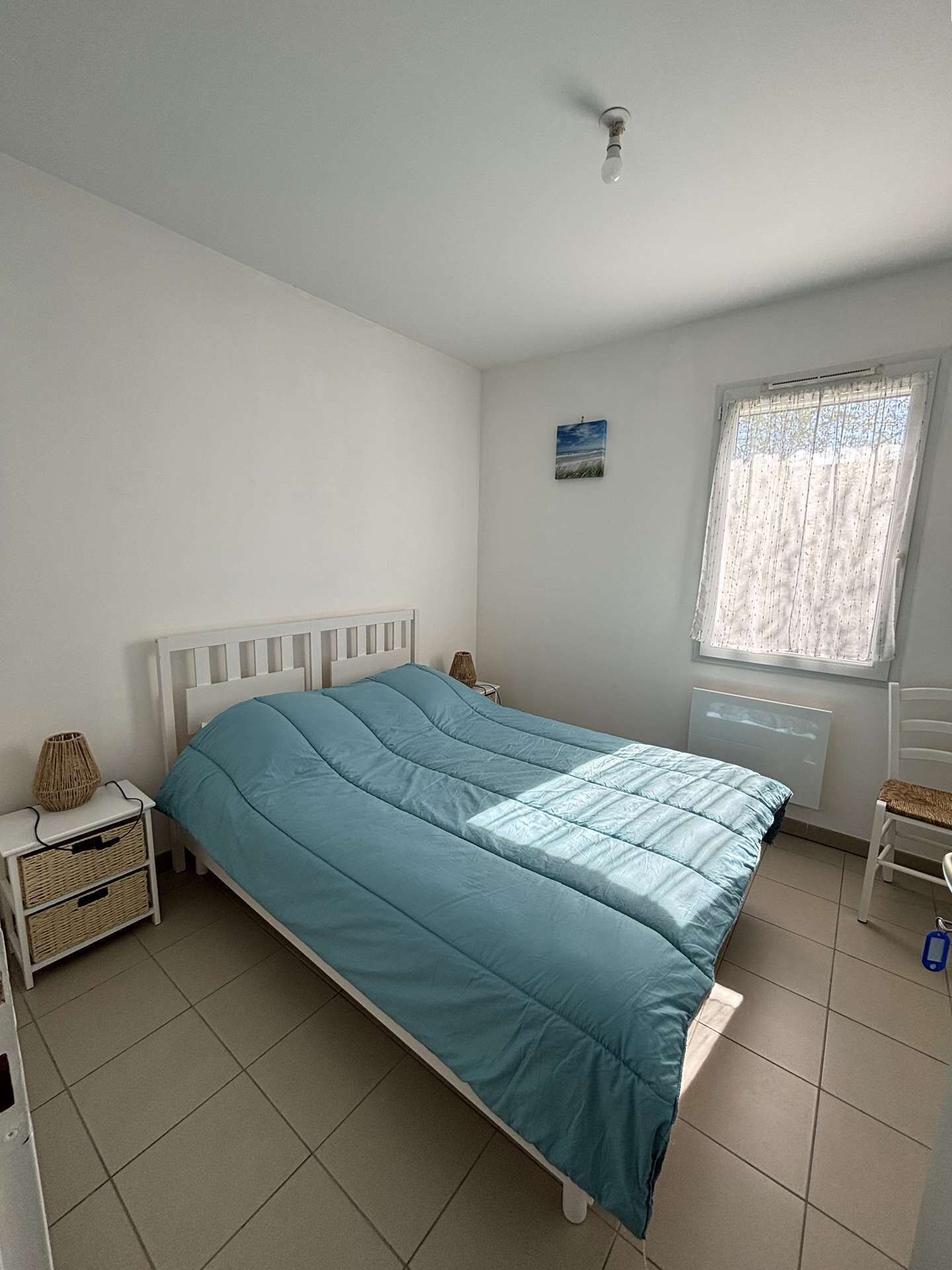 A bedroom with a bed, a nightstand, a chair, a window with curtains, a radiator, and a small wall art piece.