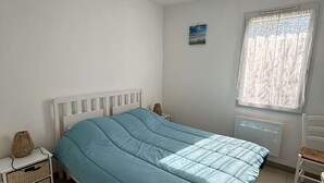Ein Schlafzimmer mit Bett, Nachttisch, Stuhl, einem Fenster mit Vorhängen, einem Heizkörper und einem kleinen Wandbild.