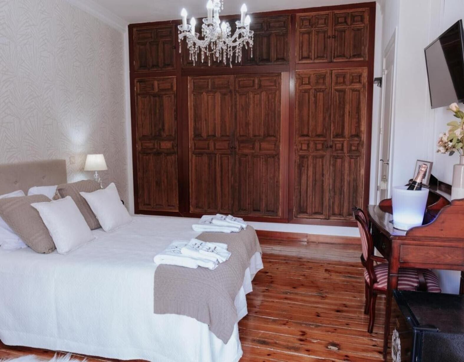 Un dormitorio con una cama grande, un armario de madera, una araña, una mesita de noche y un escritorio con una silla.