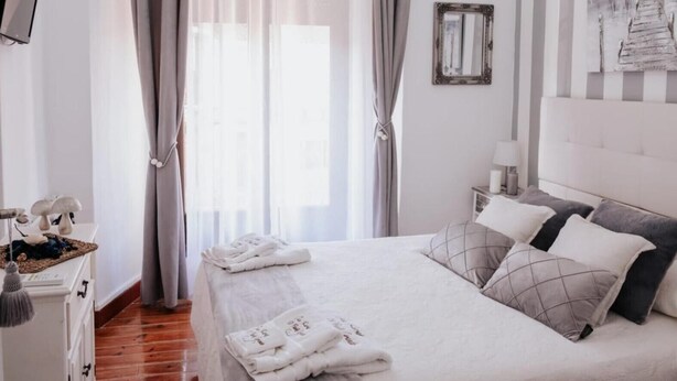 Een keurig opgemaakt slaapkamer met een bed, gordijnen, een nachtkastje met een lamp en een spiegel aan de muur.