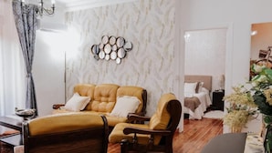 Een woonkamer met een bank, fauteuils, een kroonluchter, een spiegel aan de muur en een bed op de achtergrond.