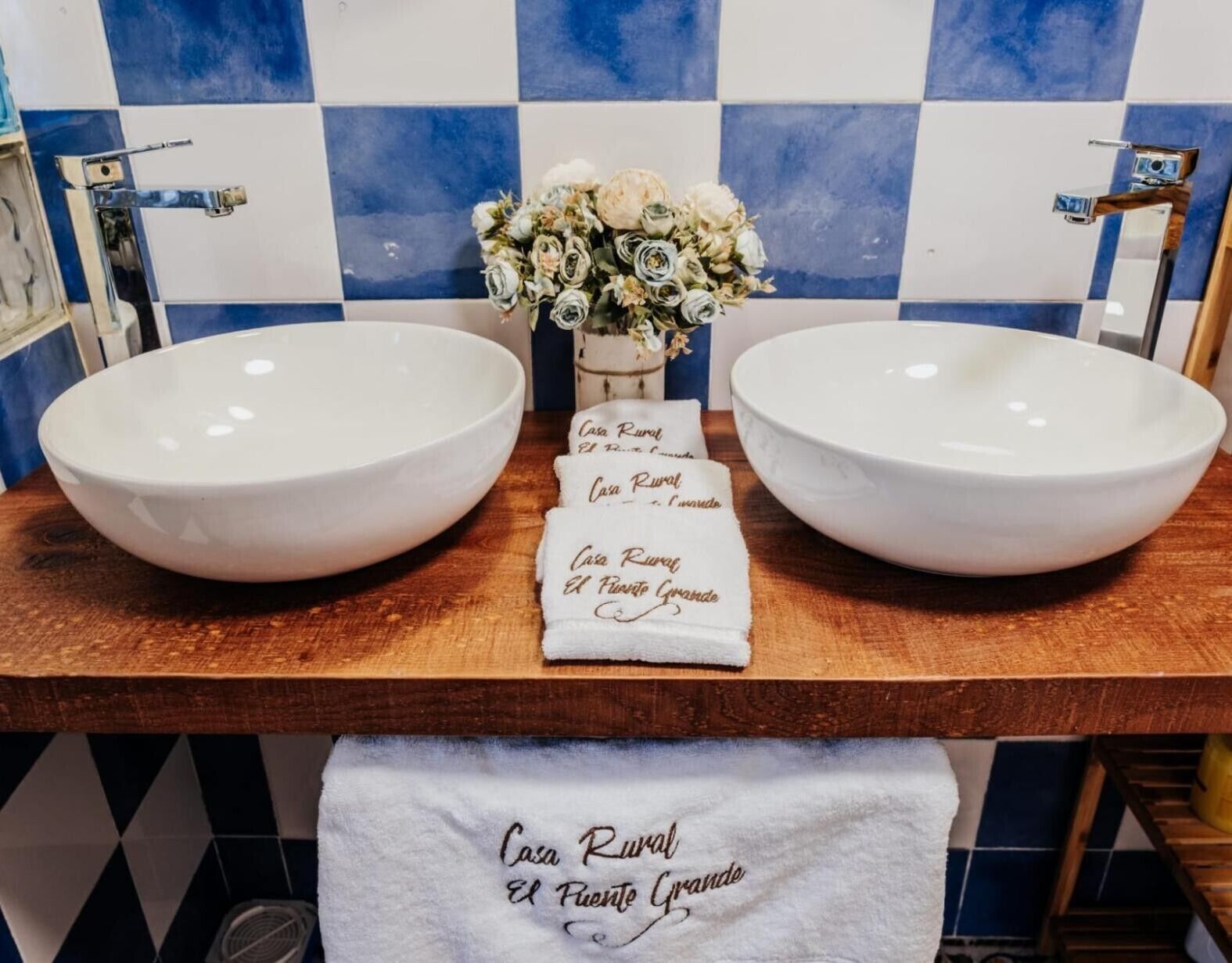 Un baño con dos lavamanos grandes de color blanco, un jarrón con flores blancas y una toalla con texto.