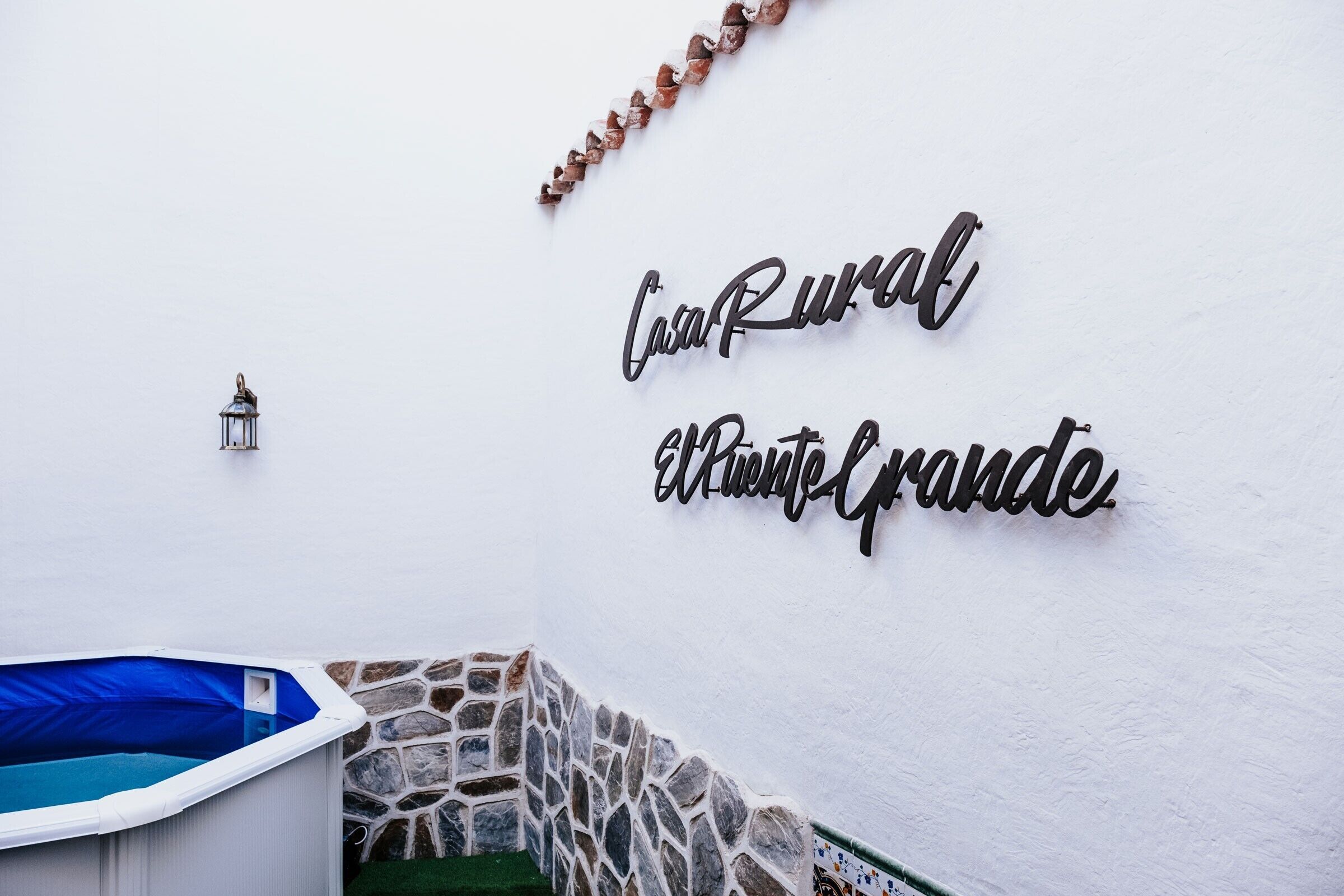 Een witte muur met een metalen bord waarop 'Casa Rural El Cuero Grande' staat en een kleine lantaarn.