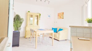 Un salon lumineux avec une table à manger, des chaises, un buffet jaune, un canapé avec des coussins bleus et une plante en pot.