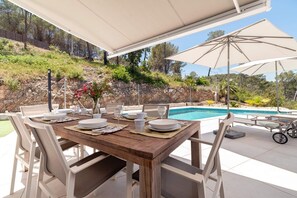 Area pranzo all'aperto con piscina, tavolo in legno, sedie bianche e ombrelloni.