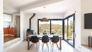 Une cuisine moderne dotée d’un grand îlot central, d’un coin repas avec des chaises et offrant une vue sur un paysage vallonné grâce à de grandes fenêtres.