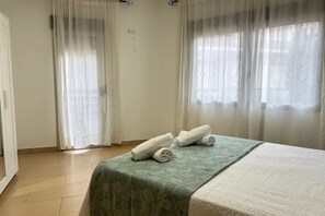 Ein Hotelzimmer mit einem Bett, einer grün gemusterten Tagesdecke, weißen Vorhängen und Blick aus dem Fenster.