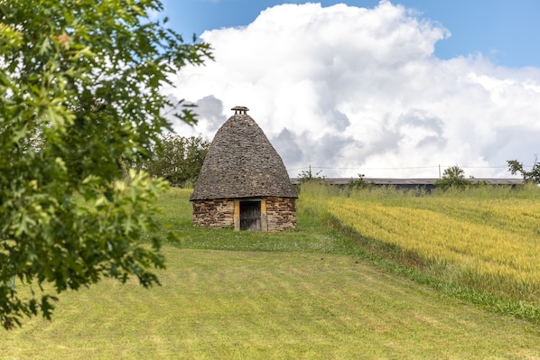 Une petite maison en pierre avec un toit conique, entourée d’un champ herbeux et d’arbres.