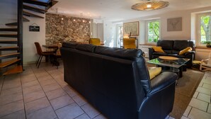 A modern living room with a black leather sofa, a dining area with chairs, a stone wall, and a staircase.