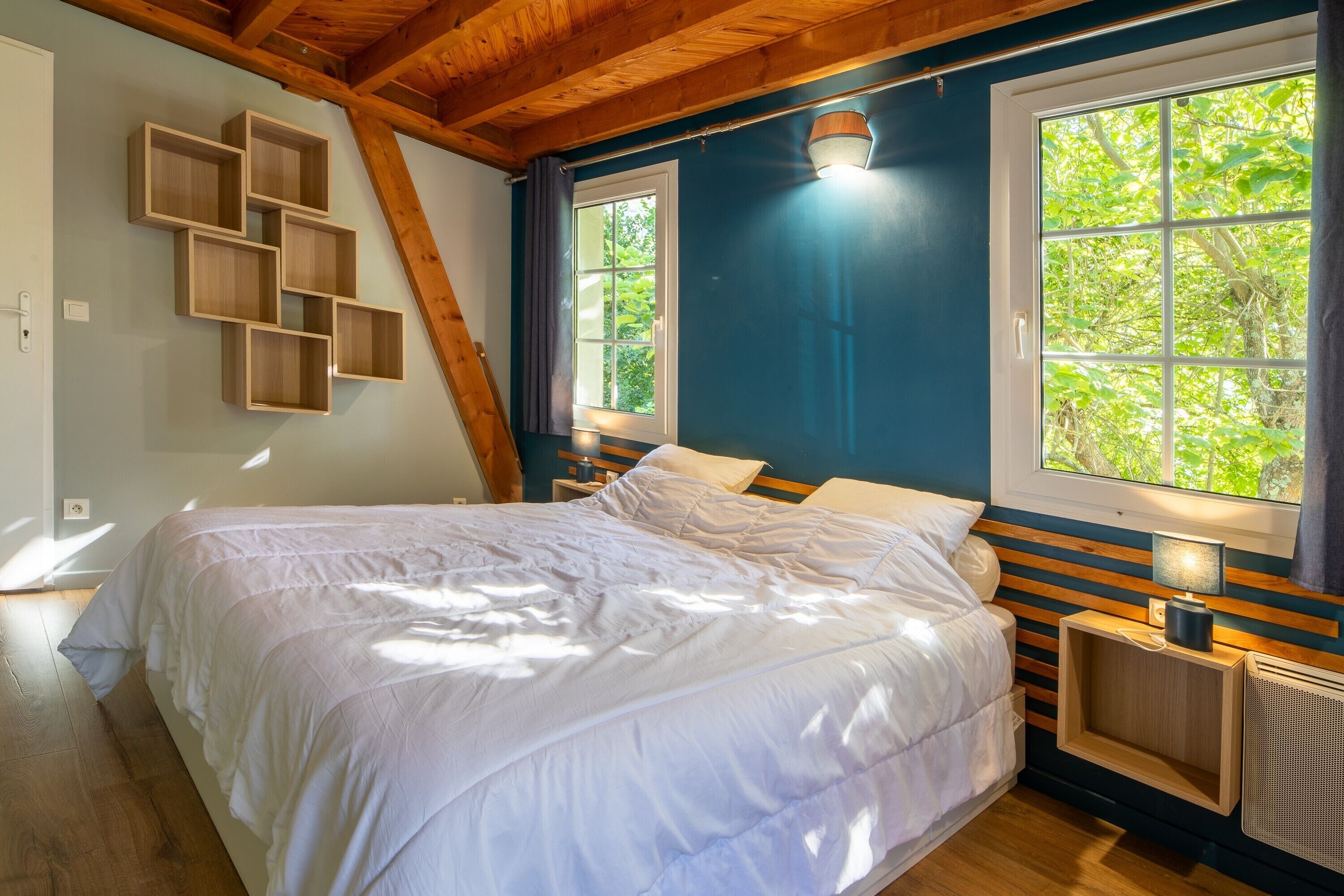 Une chambre à coucher comprenant un lit, une tête de lit en bois, deux tables de chevet, une fenêtre donnant sur un paysage verdoyant et un plafond en pente.