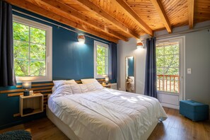 Une chambre avec un grand lit, des poutres apparentes en bois au plafond, deux tables de chevet avec des lampes, une fenêtre avec des rideaux et une porte-fenêtre donnant sur un balcon.