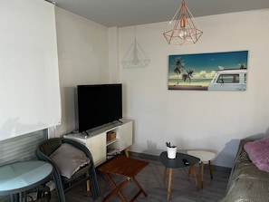 Ein gemütlicher Wohnraum mit Flachbildfernseher, einem kleinen Esstisch, einer Couch und einem Gemälde mit Strandmotiv.