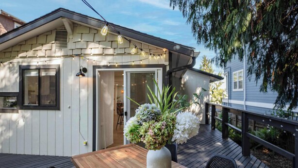 A wooden deck with a table and chairs, a white vase with flowers, and a house with a shingled roof and a door. - Stunning 4BR Modern Gem: Near UW and Green Lake! (Seattle)