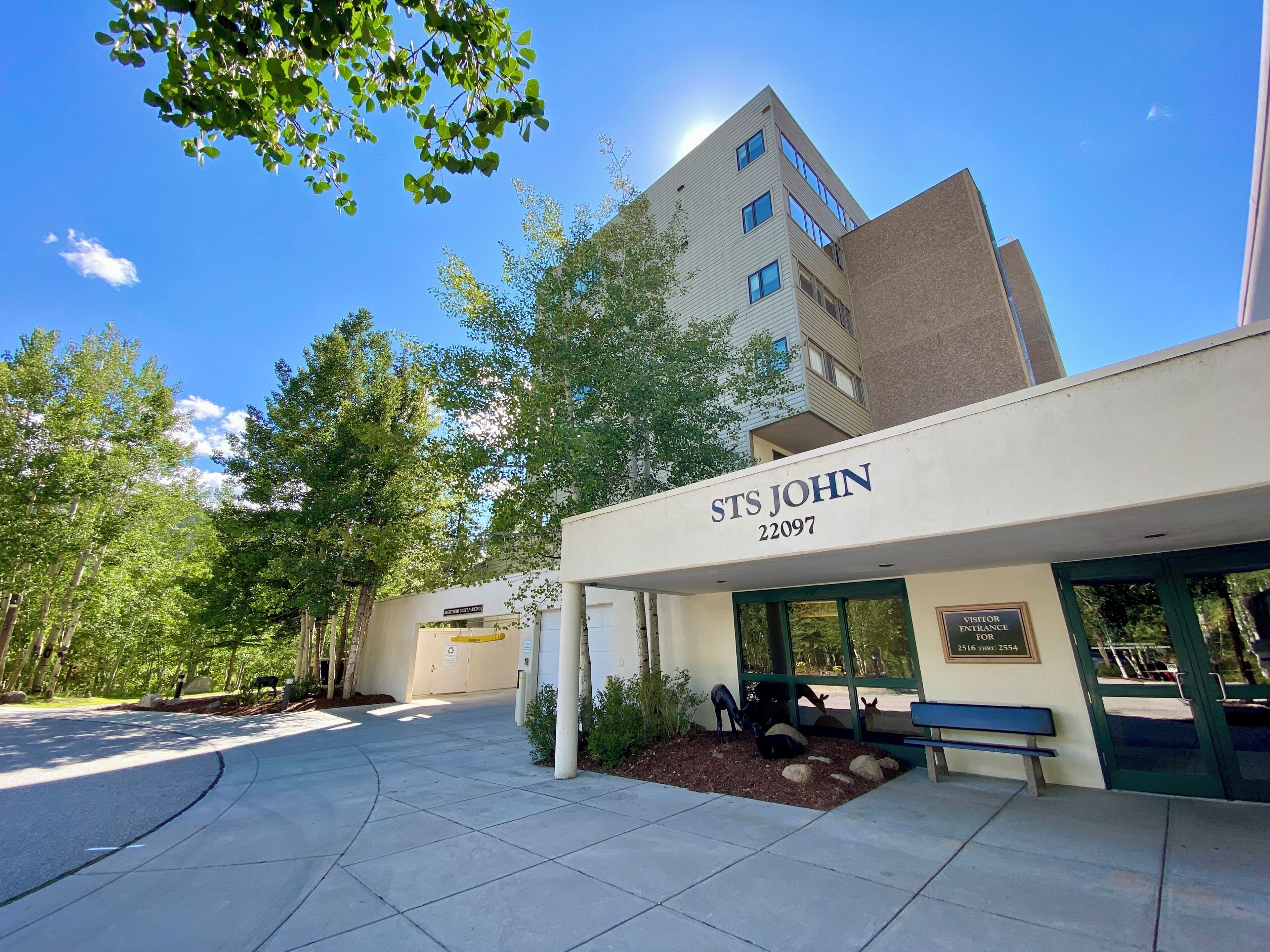 A building labeled "STS JOHN 22097" with a visitor entrance, a bench, and a paved area with trees and shrubs.