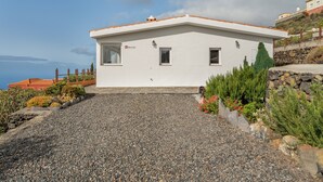 A modern white building with a sloped roof, a gravel driveway, and a stone wall with greenery.