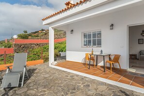 Eine überdachte Terrasse im Freien mit Tisch und Stühlen, einem Steinboden und Blick auf eine Hügelkuppe.