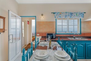 Un coin repas avec une table dressée pour quatre personnes, une cuisine avec des meubles bleus, une fenêtre donnant sur un beau paysage et une porte menant à une autre pièce.