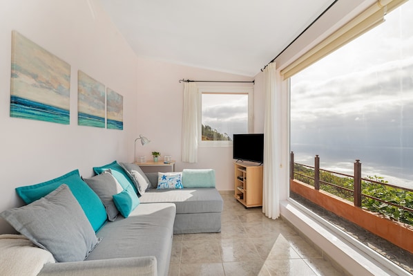 Un salon avec un canapé gris, des coussins turquoise, un téléviseur à écran plat et un balcon offrant une vue sur l’océan.