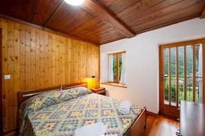 A bedroom with wooden walls, a bed with a quilt, a nightstand, a window, and a balcony view. - Apartment 'Luéisch Ketschu 2' with Mountain View, Shared Garden and Wi-Fi (Issime)