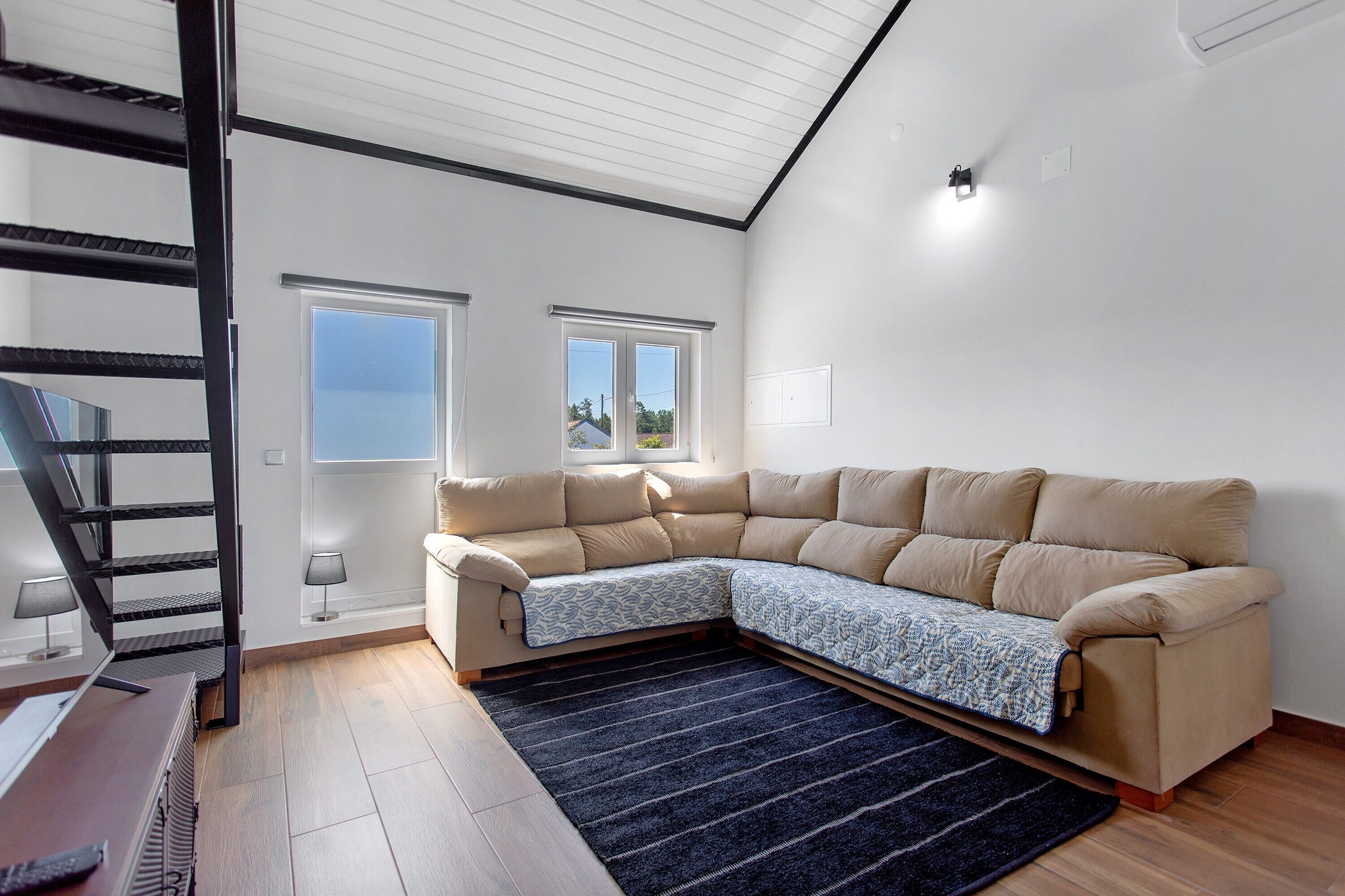 Un salon moderne avec un canapé d’angle, un escalier et deux fenêtres.