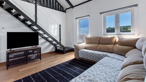 Una sala de estar moderna con una escalera, un sofá, un televisor sobre un mueble bajo y ventanas amplias.
