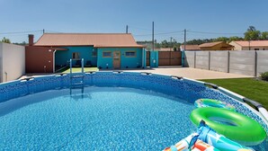 Una piscina con flotadores y una escalera, rodeada por una casa de techo rojo y un patio cercado.