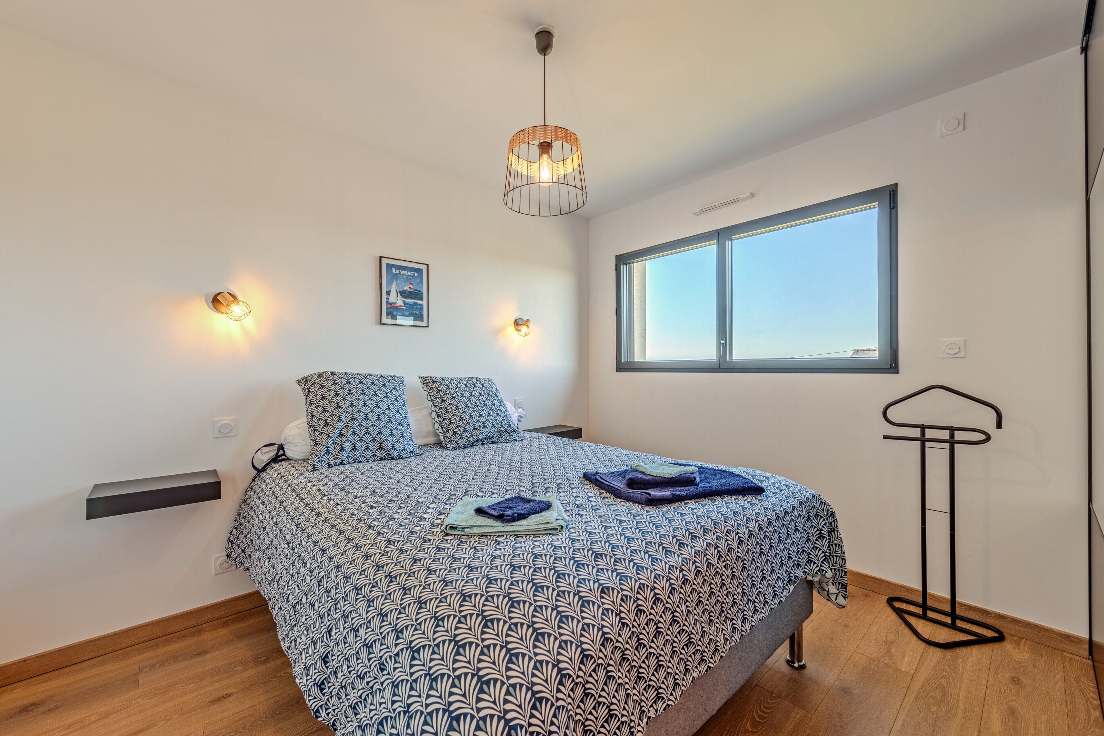 Ein Schlafzimmer mit einem gemusterten Bettüberwurf, einer Hängelampe, einem Fenster und einem an der Wand befestigten Regal.