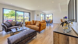 Un amplio salón con sofá, reposapiés y mesa de centro, ventanas grandes y un comedor con sillas y mesa.