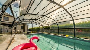 Una piscina con cerramiento de vidrio, un flotador de flamenco rosa y vistas a una casa y vegetación.