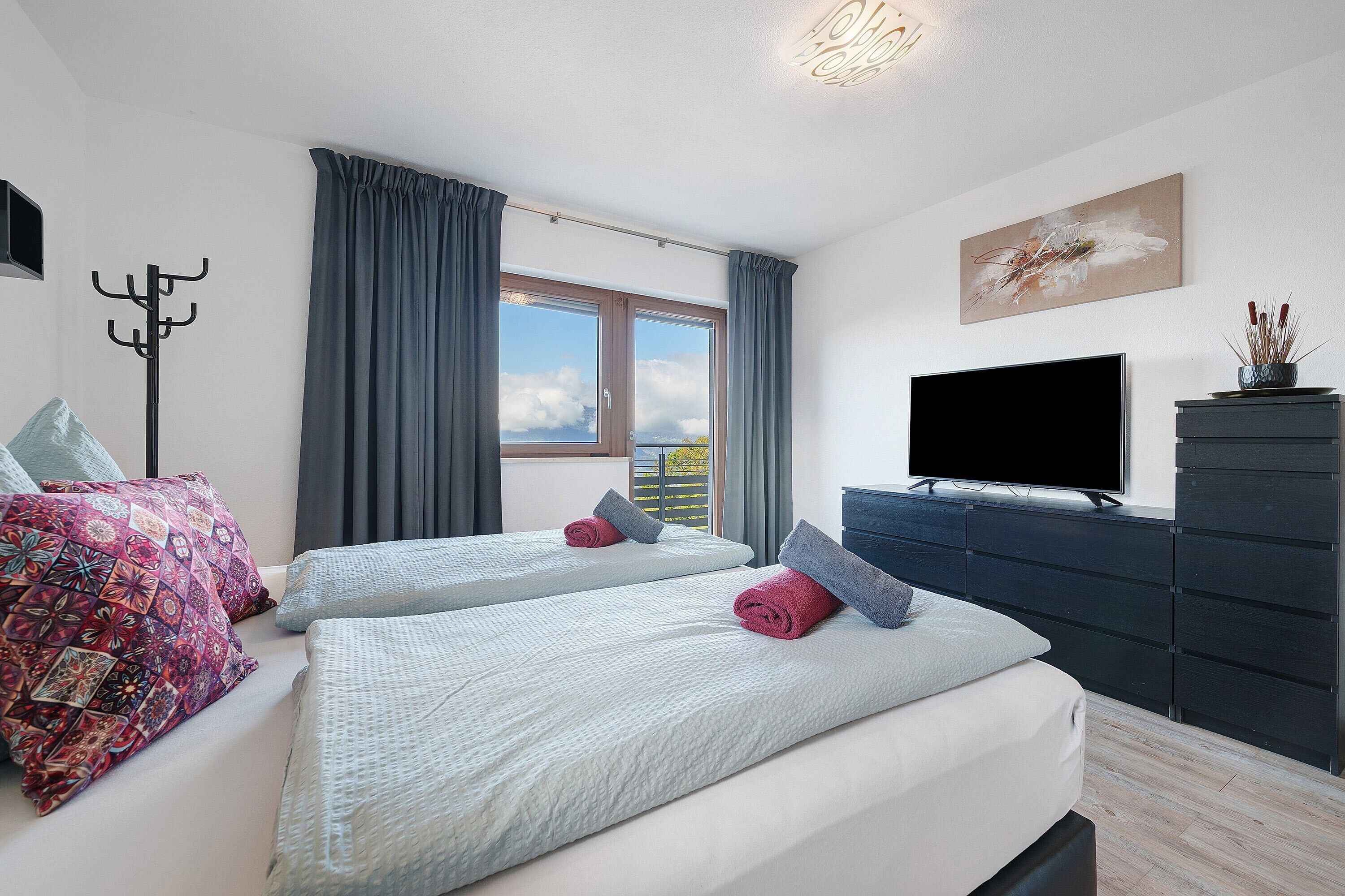 A bedroom with two beds, a TV, a coat rack, and a window with curtains.