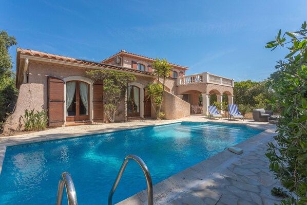 A poolside view of a house with a swimming pool, a covered patio, and a stone pathway.
