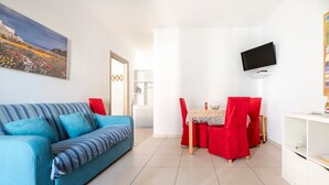 Una sala de estar con un sofá, una mesa de comedor con sillas rojas, un televisor montado en la pared y una estantería.