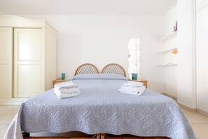 Uma cama bem arrumada com um edredom cinza, duas cabeceiras de madeira, uma mesa de cabeceira com abajur e um guarda-roupa branco.