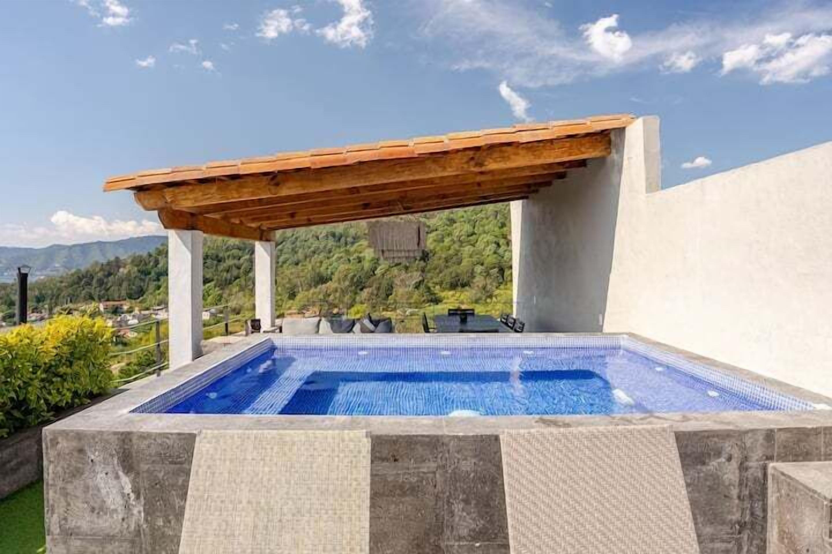 A rectangular outdoor pool with a wooden pergola overhead, surrounded by a low stone wall and greenery.