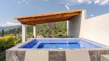 A rectangular outdoor pool with a wooden pergola overhead, surrounded by a low stone wall and greenery.