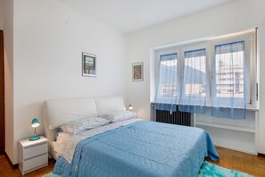 Un dormitorio con cama, mesita de noche, lámpara, ventana con cortinas y un radiador.