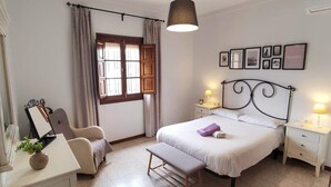Un dormitorio con cama, mesitas de noche, escritorio, silla, ventana con cortinas y cuadros en la pared.