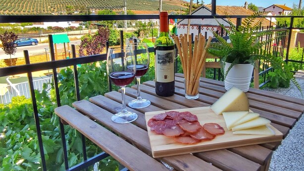 Una mesa de madera con una botella de vino, copas, una tabla de quesos y palitos de pan en un balcón con vistas a un viñedo y casas.