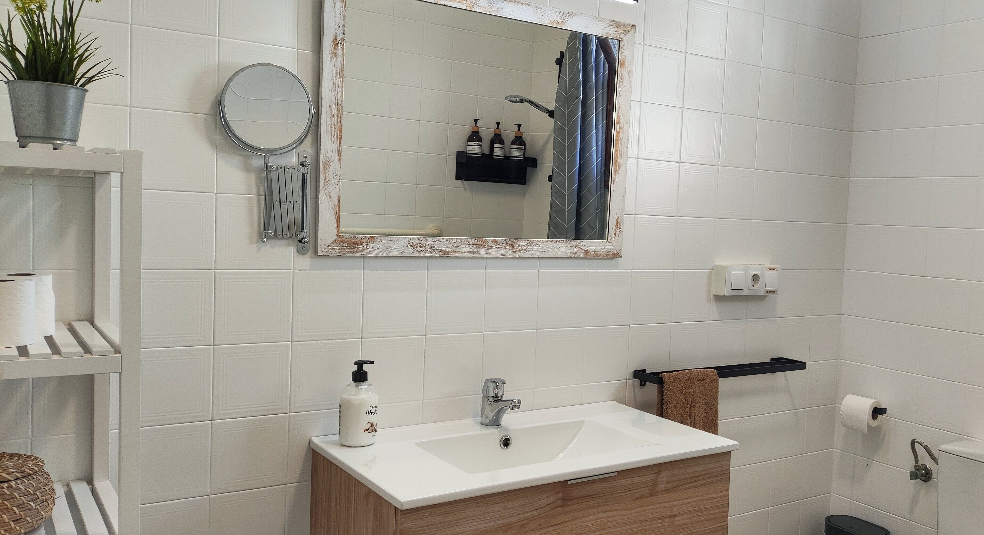 Un baño con un lavabo blanco, un espejo, un perchero para toallas y un estante con artículos de aseo.