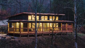 Una cabaña con terraza de madera y ventanas amplias, rodeada de árboles.