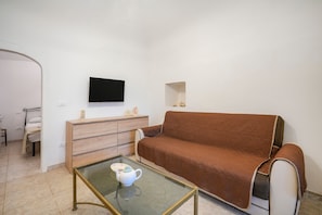 Ett rum med en brun soffa, ett glasbord med en tekanna, en byrå i trä, en platt-TV som är monterad på väggen och en liten hylla med dekorativa föremål.