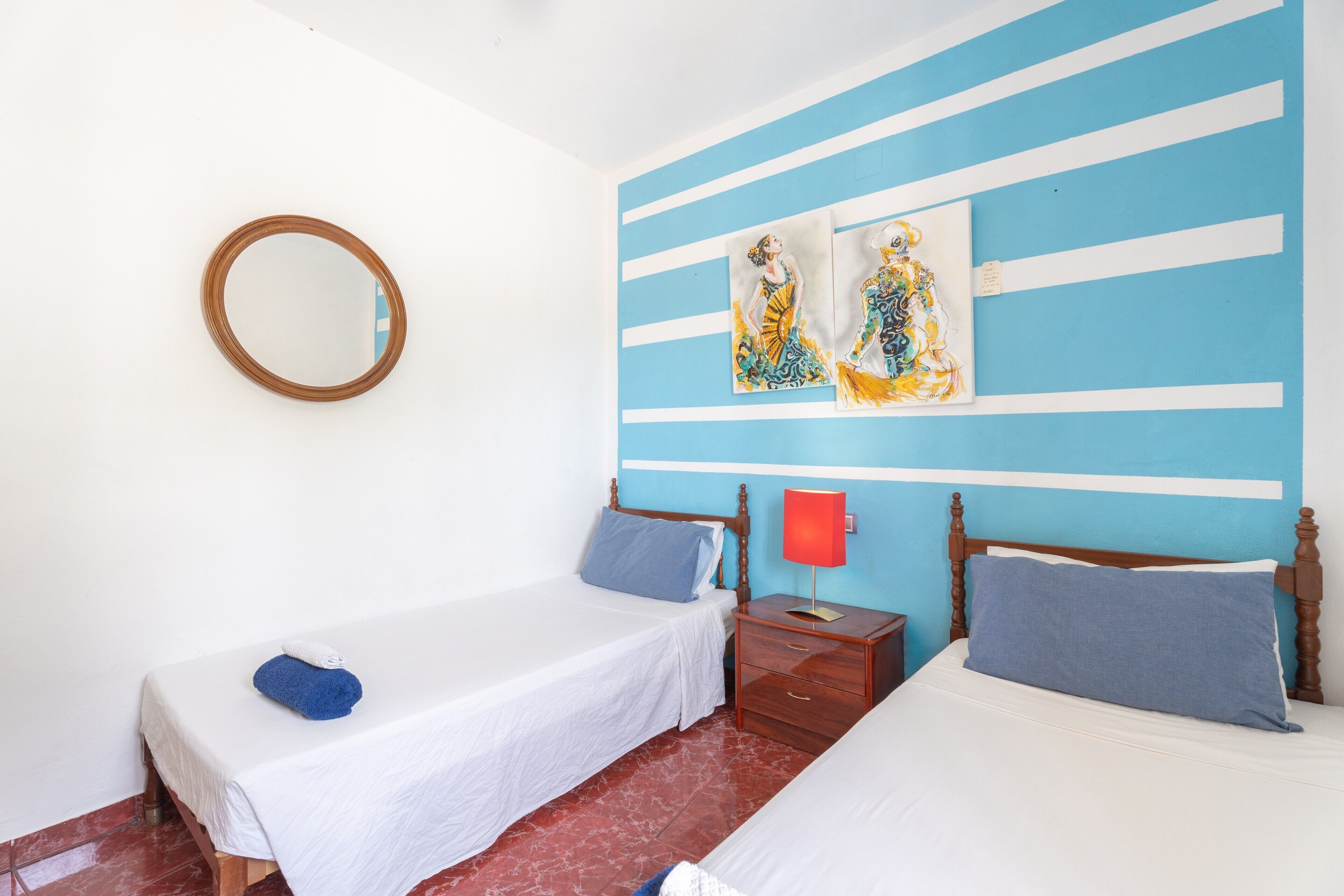 Una habitación con dos camas, una mesita de noche de madera, una lámpara roja y un espejo en la pared.