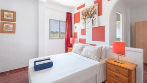 Een slaapkamer met een bed, een nachtkastje, een lamp, een plafondventilator, een raam met gordijnen en kunst aan de muur.