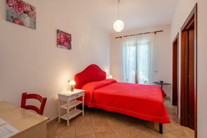 Une chambre à coucher comprenant un lit, un bureau, une chaise, une table de chevet, une lampe, une fenêtre avec des rideaux et une porte.