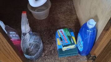 A kitchen sink with a black trash bin, a plastic bottle, a box of dish soap, and a blue detergent bottle.